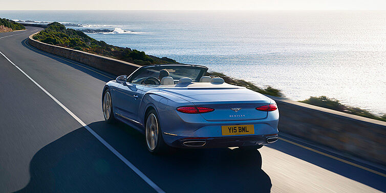 Bentley Katowice New Bentley Continental GTC Azure convertible rear view in Light Windsor blue paint with 22 inch ten spoke wheels driving dynamically on an ocean road in the sunshine